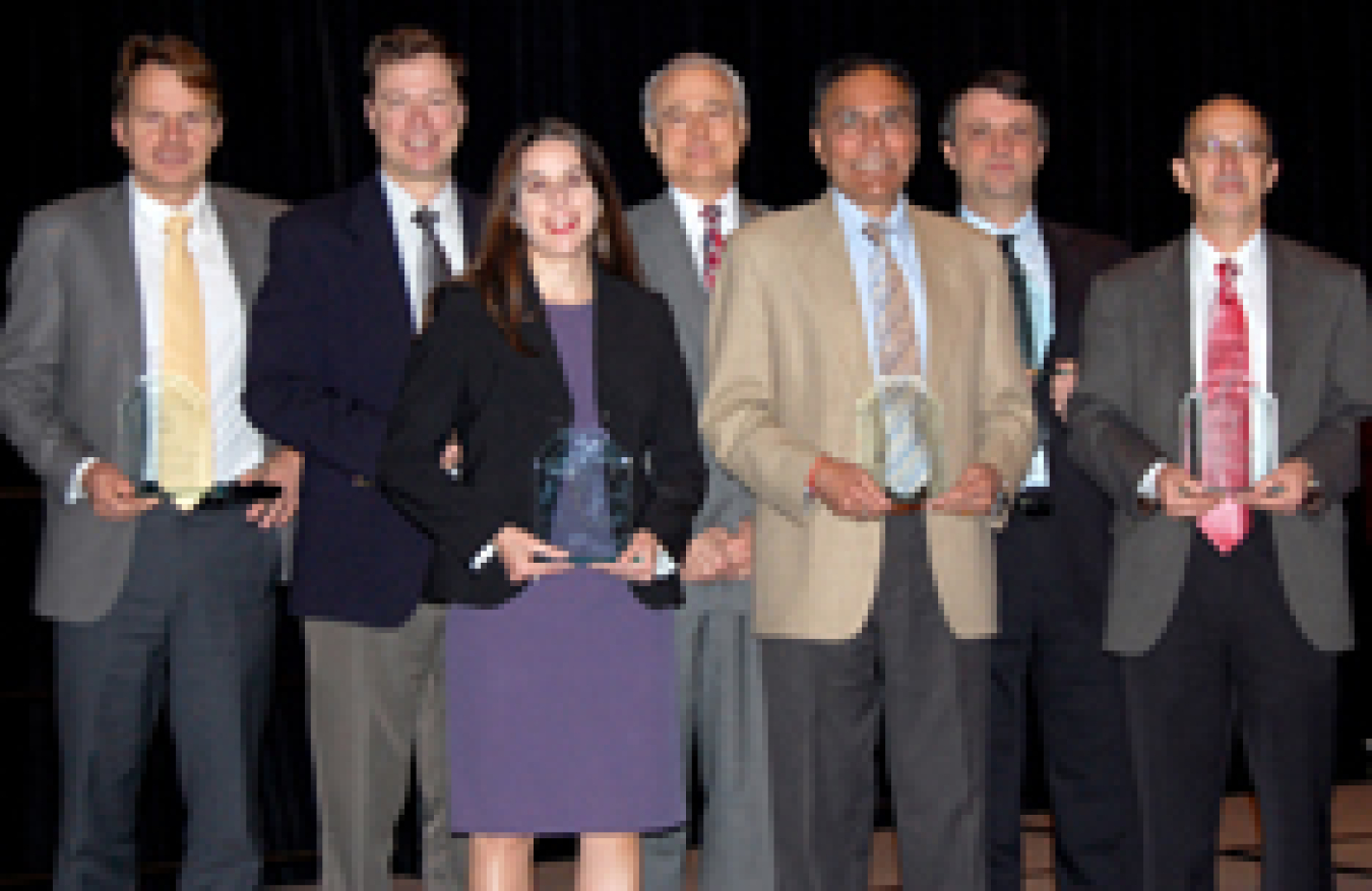 Photo of one woman and six men standing in a row facing the camera. Everyone is holding a small acrylic award except for one man in the middle of the photo.