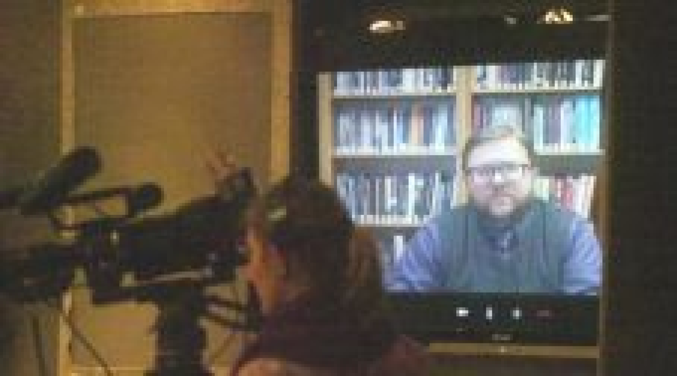 Photo of a woman with a video camera in a room set up in front of a screen showing a man facing the camera via Skype.