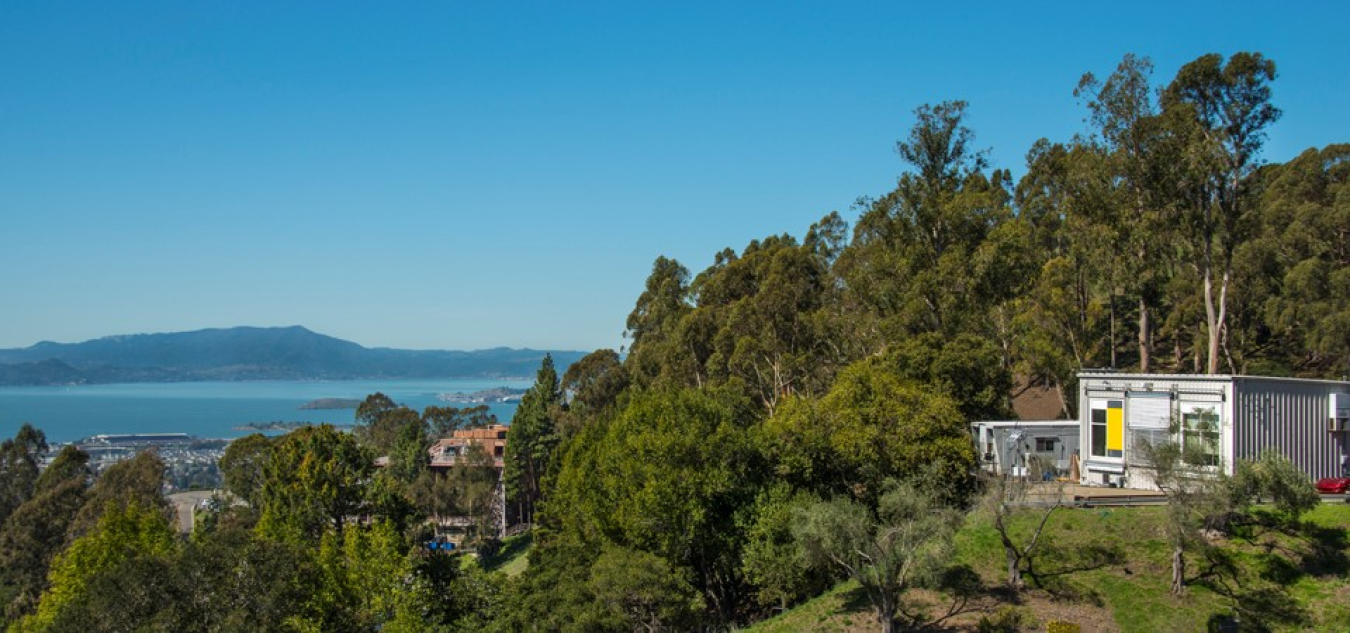 An expanse of trees and a home on a cliff, with a lake and mountains beyond.