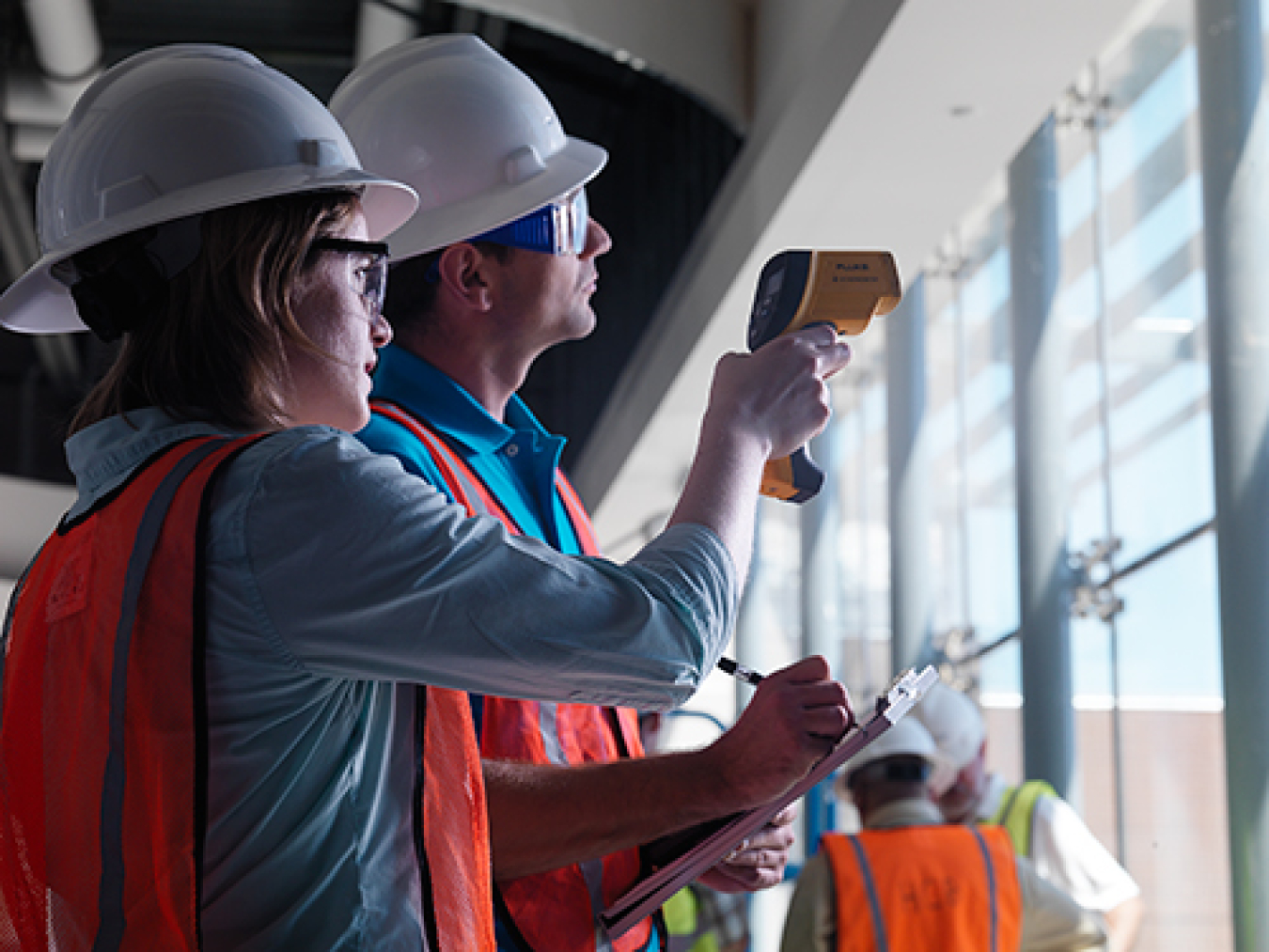 A man and a woman in hard hats working in a building, in front of windows.