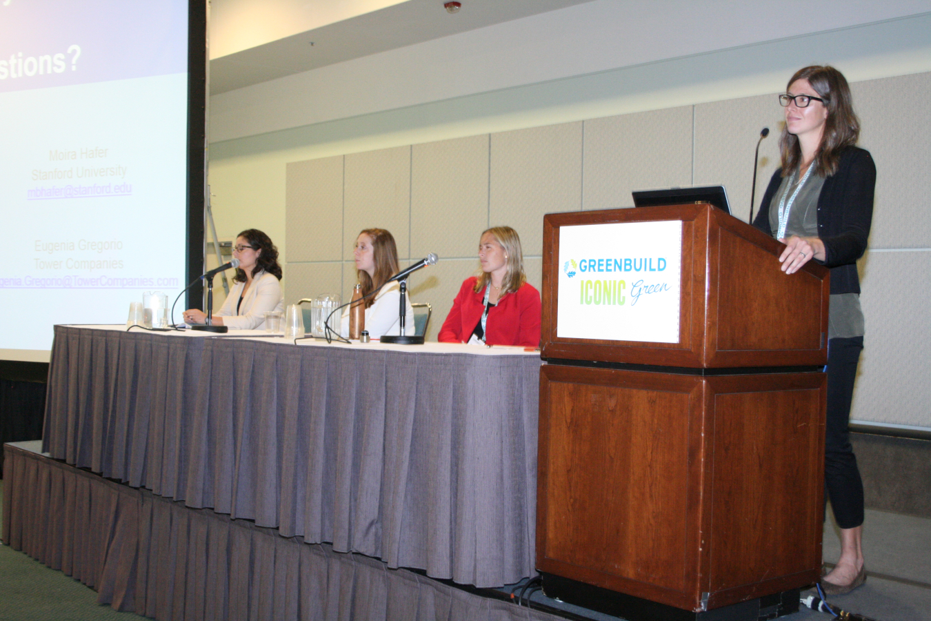 Four females addressing an audience at the front of a meeting room, with a projection screen off to the side. Three females are sitting at the long table, and the fourth is standing at a lectern.