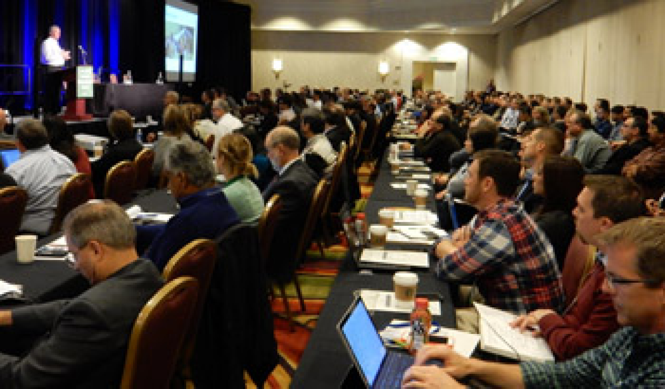 Panel attendees seated in a large room listening to a speaker, some with laptops.