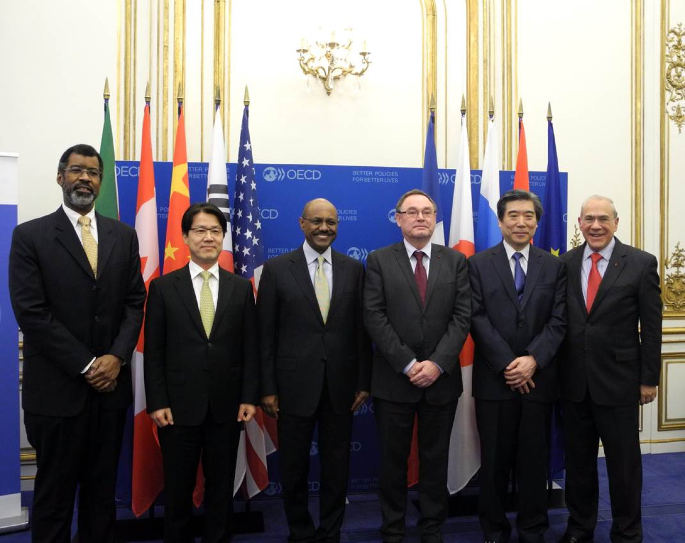 Group of six men in dark business suits and ties standing in front of a sign and a series of flags, looking at the camera.