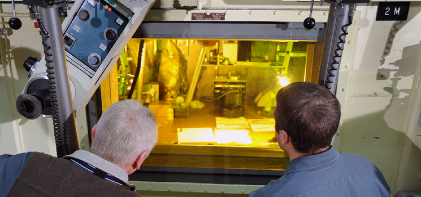 Photo of two men, backs to the camera, looking through a tinted window at a part of the fuel cycle process.