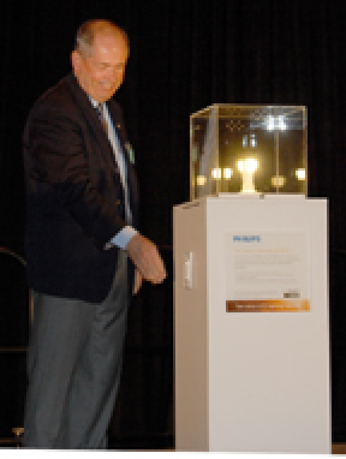 Photo of a man standing next to a pedestal which has a lighted bulb on top, covered by a clear acrylic box. The man's hand is extended toward a switch on the side of the pedestal, as if he were turning the switch on or off.