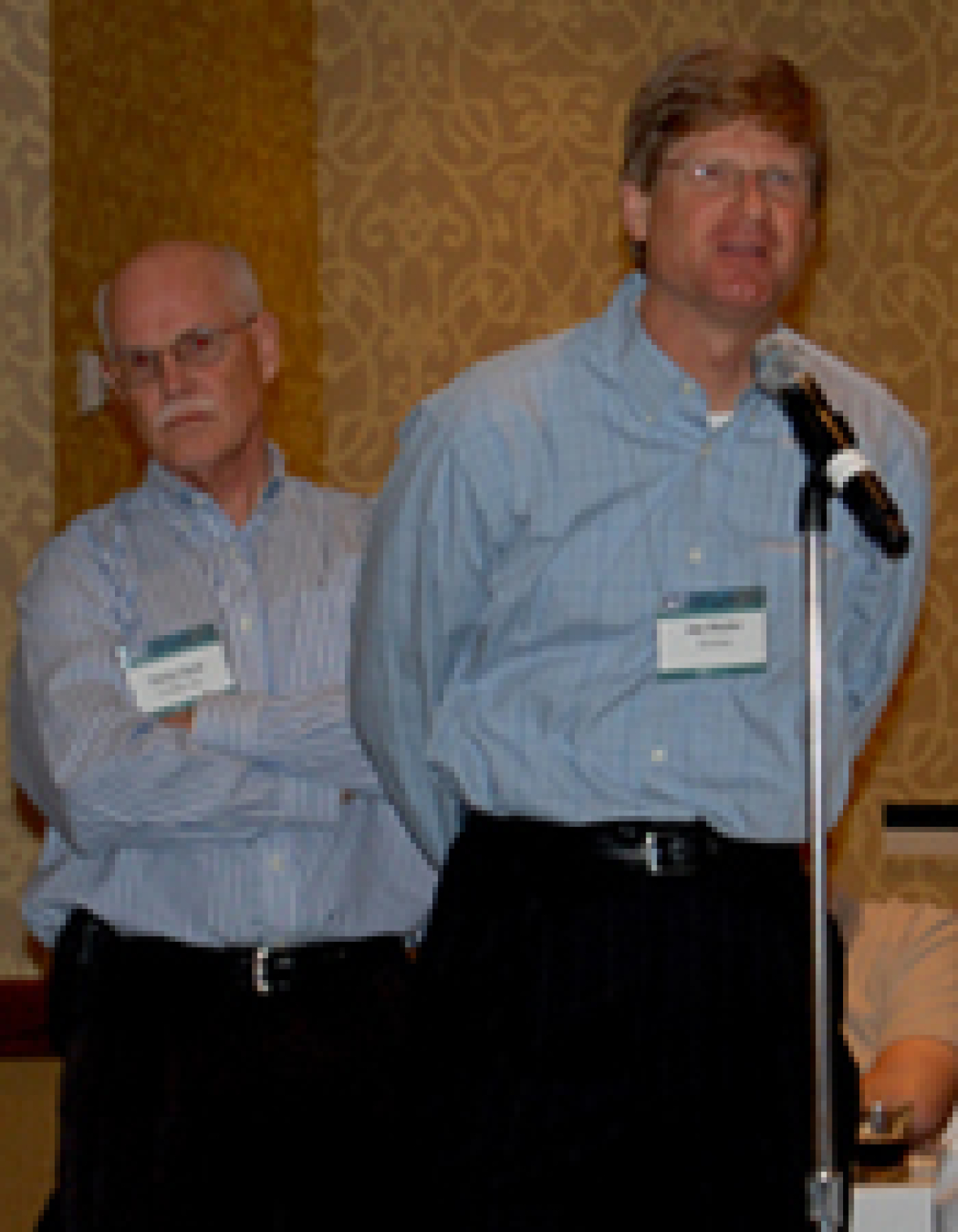 Photo of two men standing in line behind a microphone. One man is talking in the microphone as though asking a question.