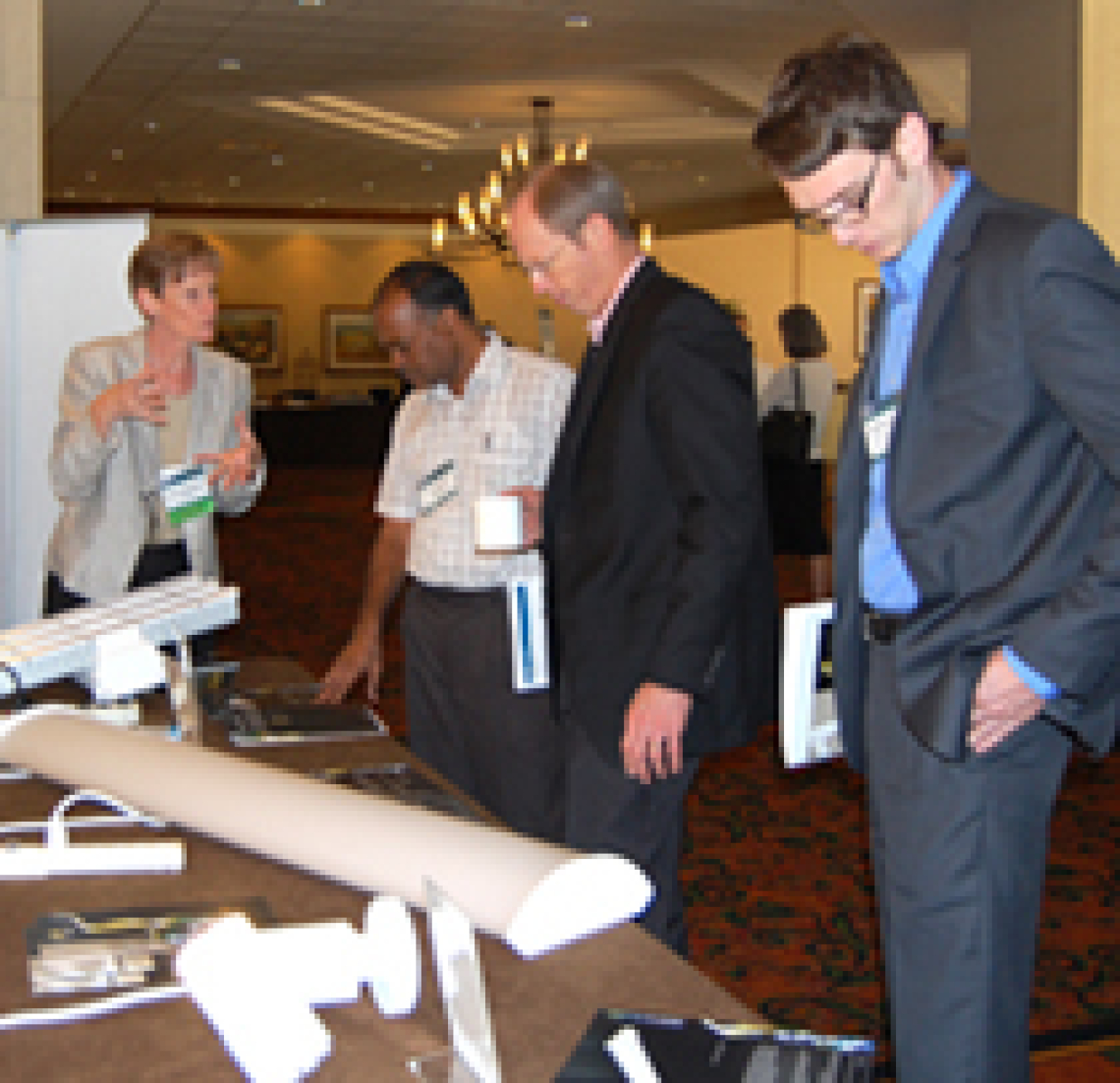 Photo of three men and one woman standing in front of a table containing assorted light fixtures. The woman is talking to the three men.