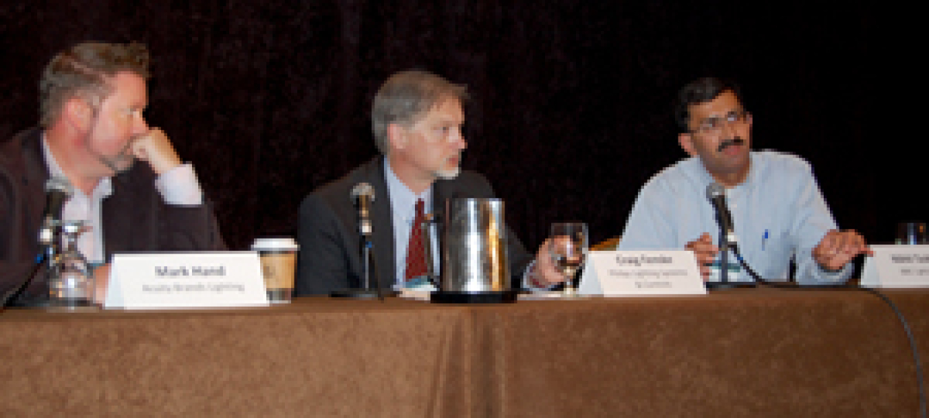 Photo of three men sitting side by side on a conference panel. Two men are looking at the third man, who is talking into a microphone.