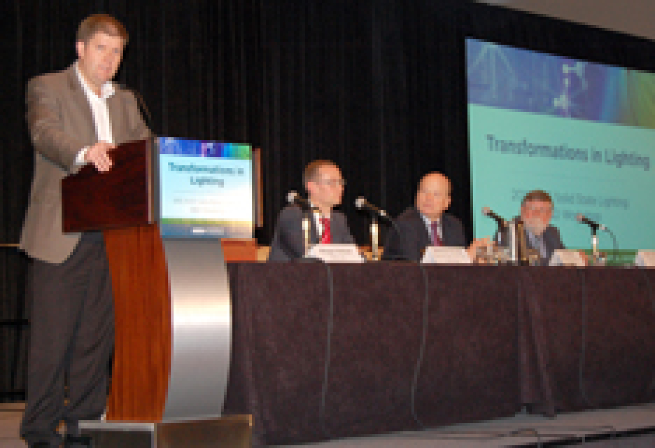 Photo of three men sitting at a table with microphones on it. Another man is standing at a podium.