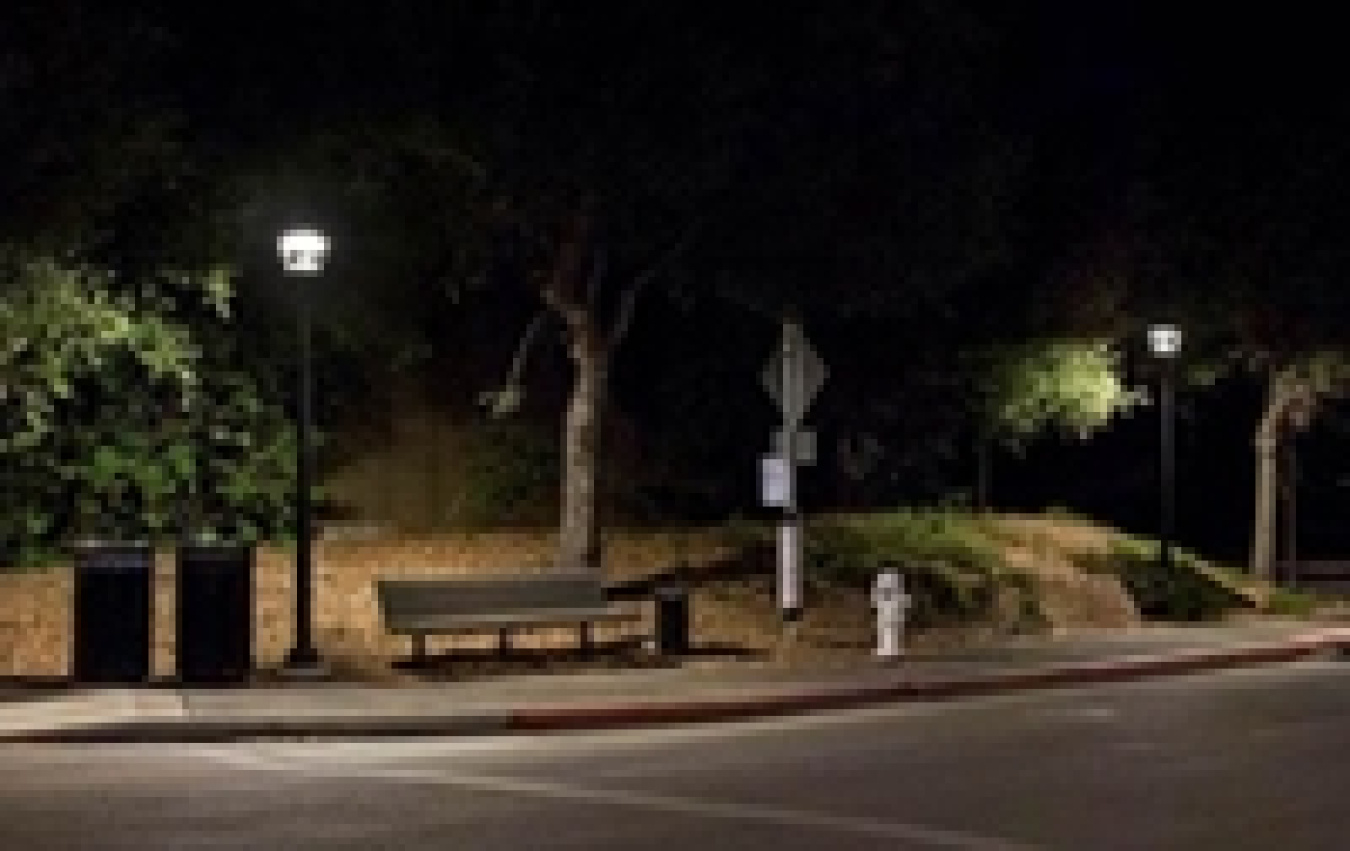 Photo of a sidewalk at night with a bench in the middle and streetlights on either side.