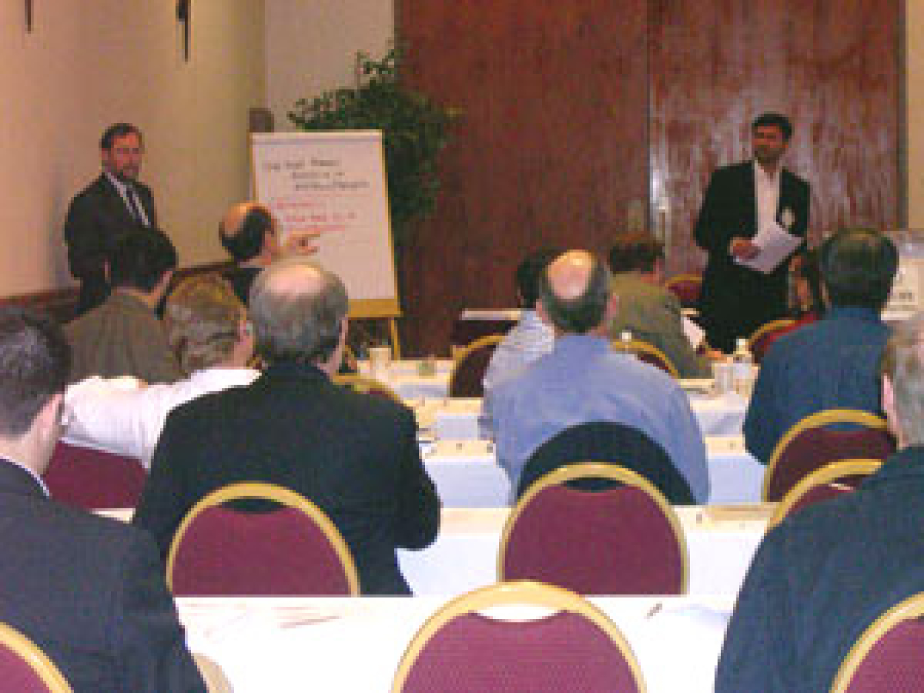 Photo taken from the back of a conference room, showing the back of people's heads, and in the front, a man standing next to an easel with paper, and to the far right a man standing with a sheet of paper in his hand.