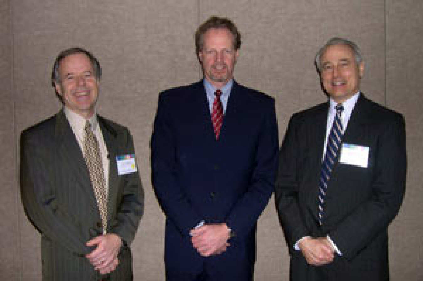 Photo of three men in suits, standing with their hands folded, smiling at the camera.