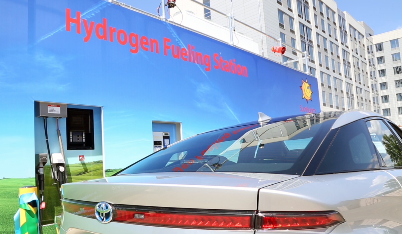 A fuel cell vehicle drives by a dmonstration building painted with the words Hydrogen Fueling Station.