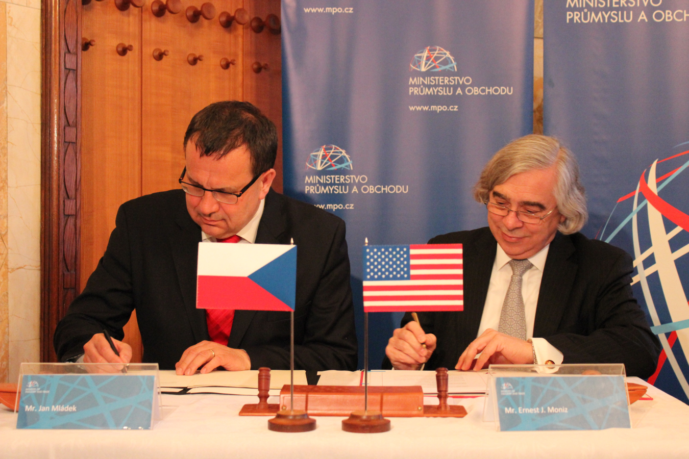 Two men seated at a table signing documents. One (Jan Mládek) has the Czech flag in front of him on the table, while the other (Secretary Ernest Moniz ) has the U.S. flag in front of him.