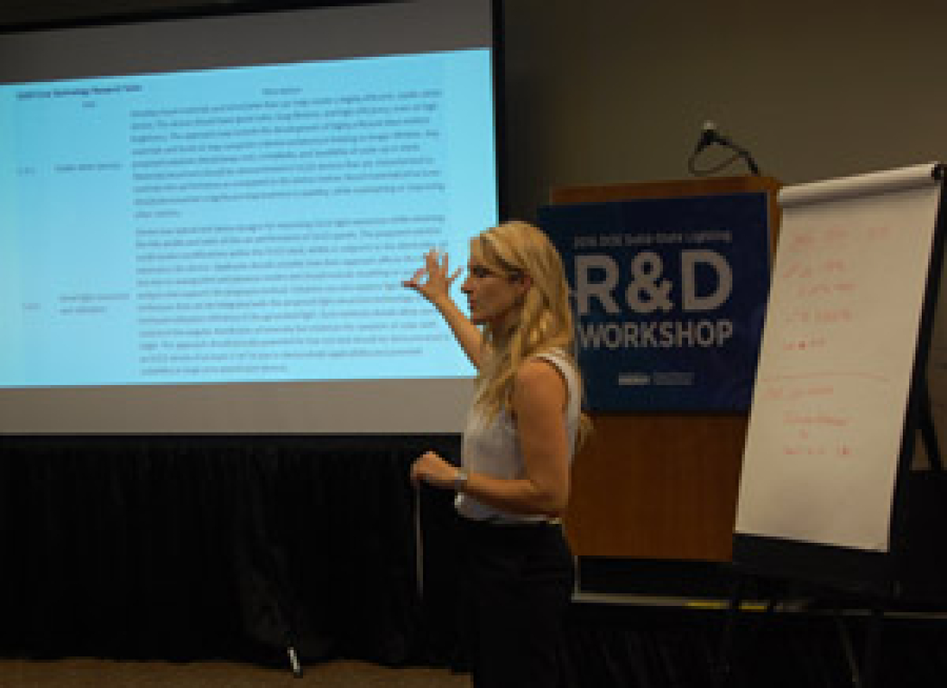 Woman speaking to an audience and motioning toward a projection screen.