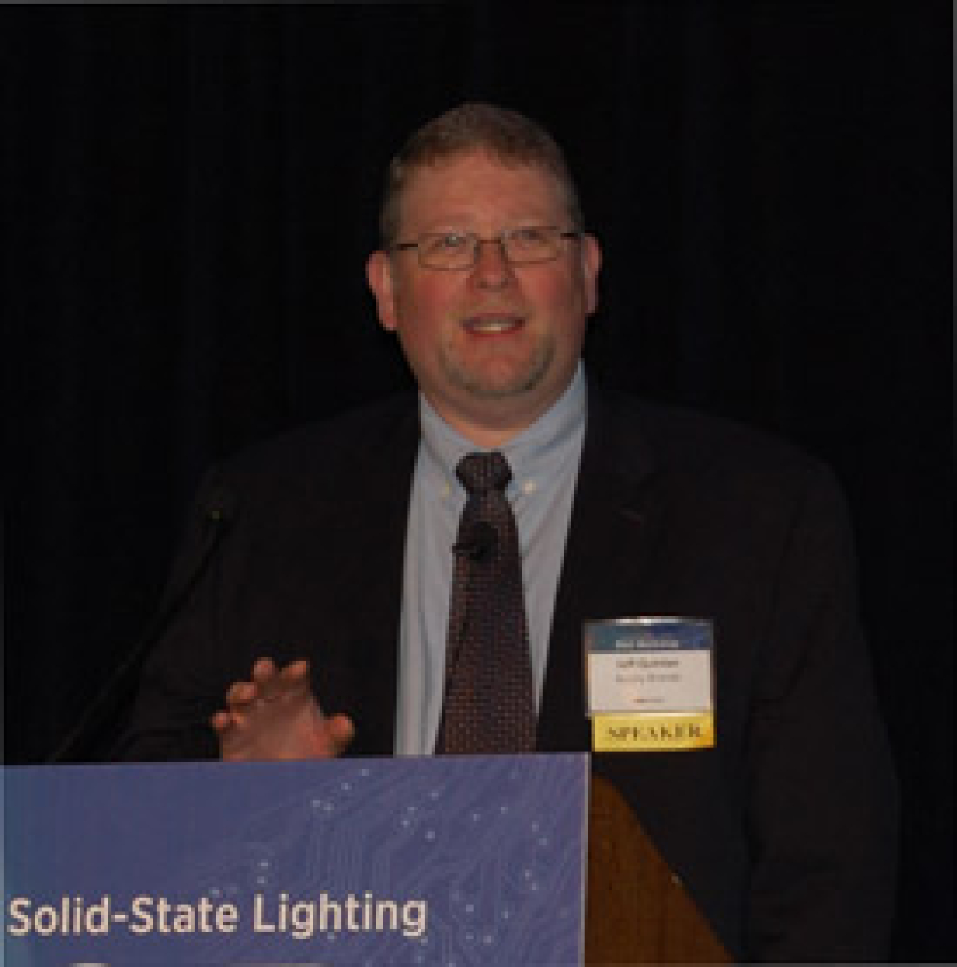 Man in a suit standing at a lectern addressing an audience.