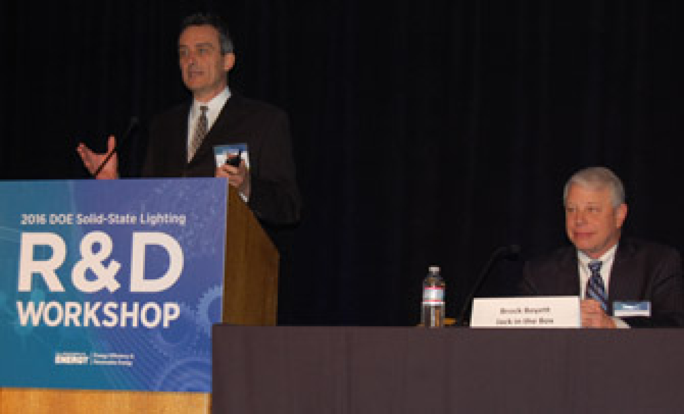 Two men, one seated on the right and another standing at a lectern on the left, addressing an audience.