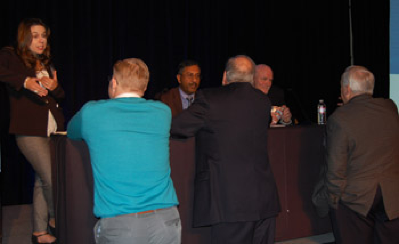 Group of people talking to each other at the front of a conference room, near a table.