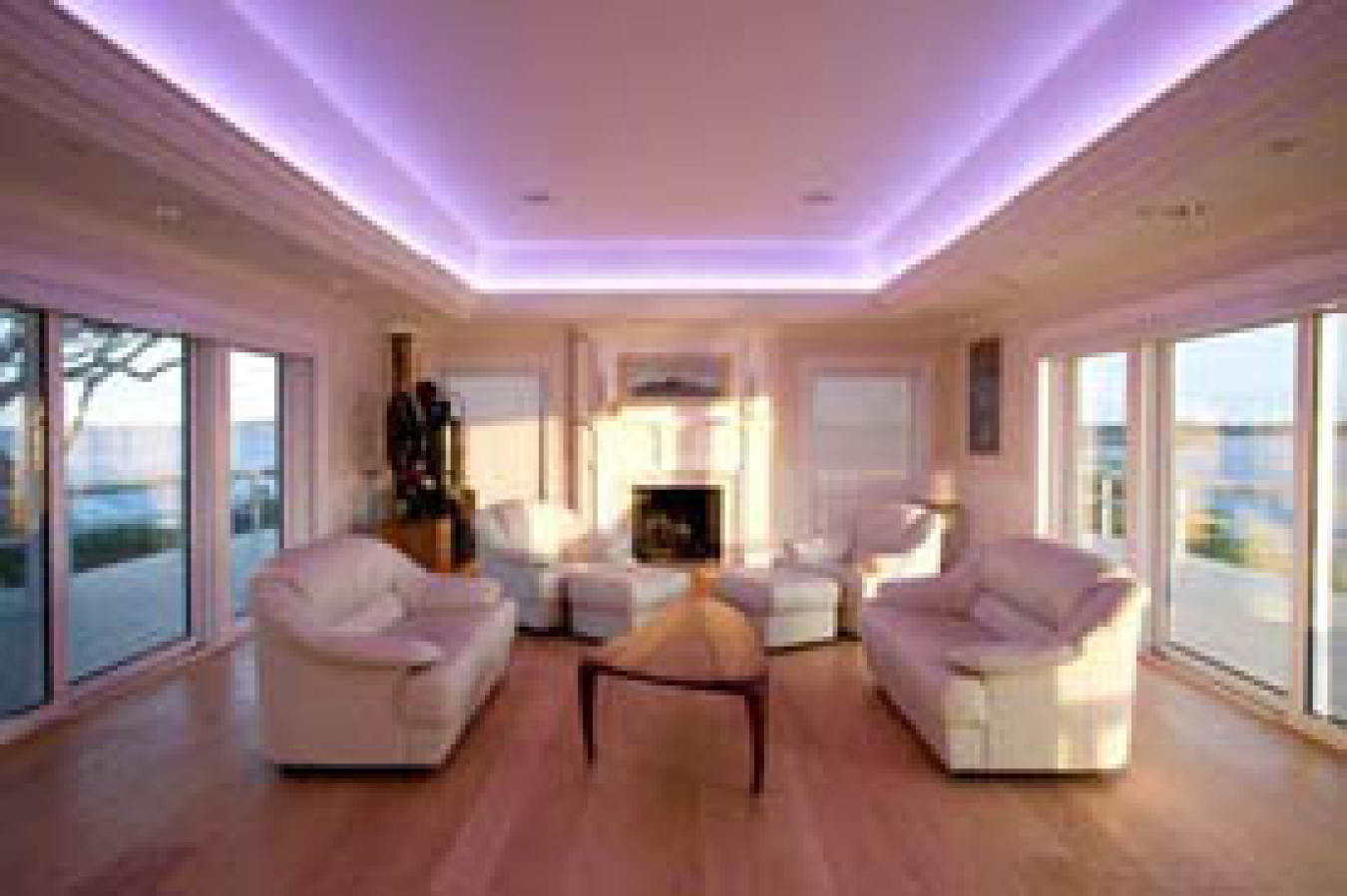 Photo of a living room with floor to ceiling windows on each side. There are two white couches facing each other in the middle of the room with a triangular coffee table between them, and the ceiling sports a glowing square of recessed ceiling lights.