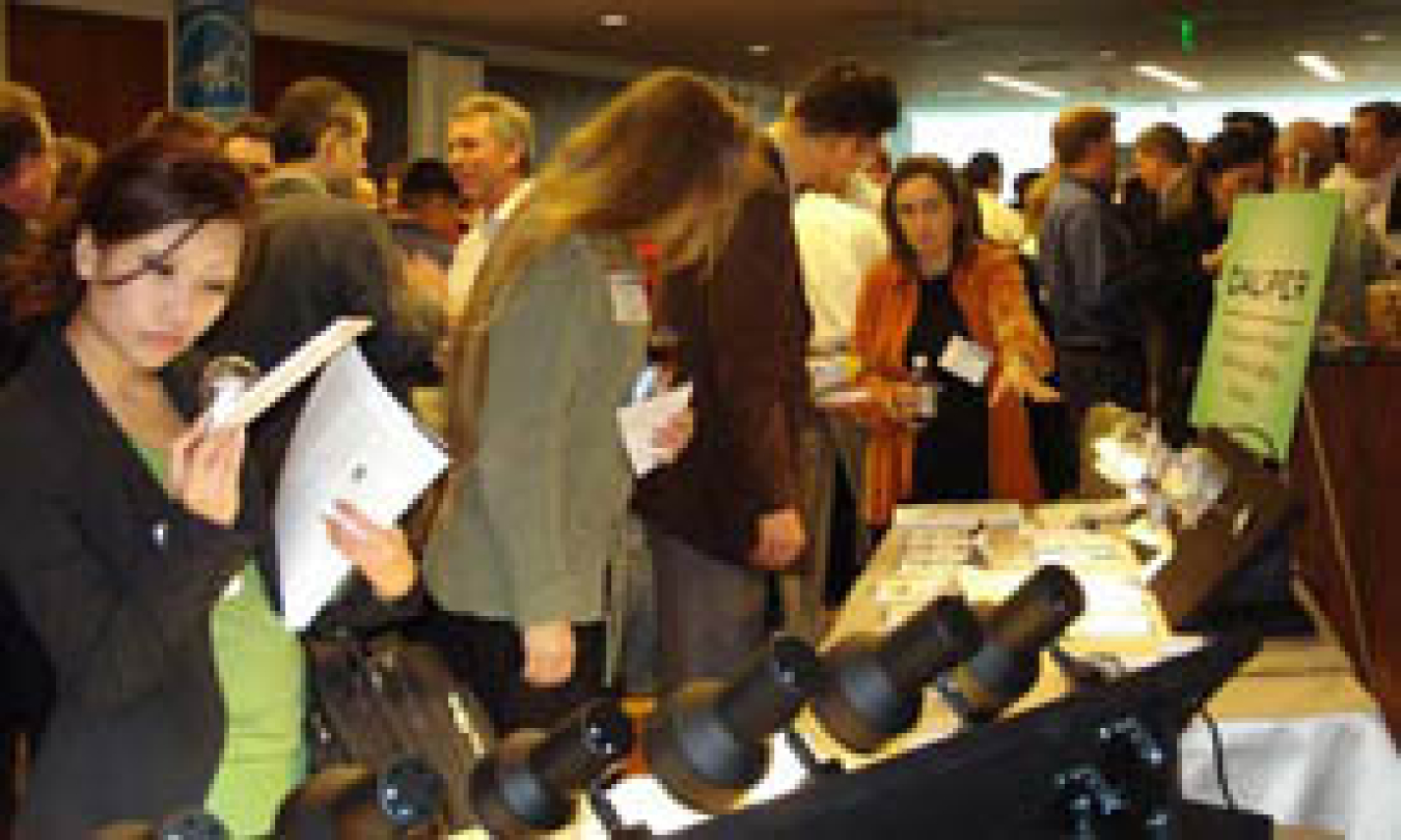 Photo of a crowd gathered around a display booth. There is an Asian woman examining some literature in the foreground.