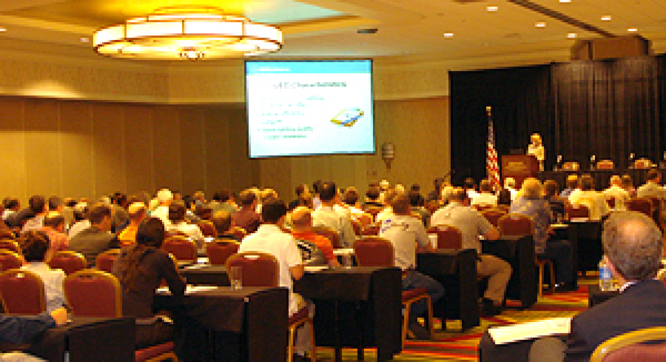 Photo taken from the back of a large ballroom showing the backs of a large crowd seated viewing a presentation on a large screen in the center background, and a speaker in the distance on the right standing at a podium next to the U.S. flag.