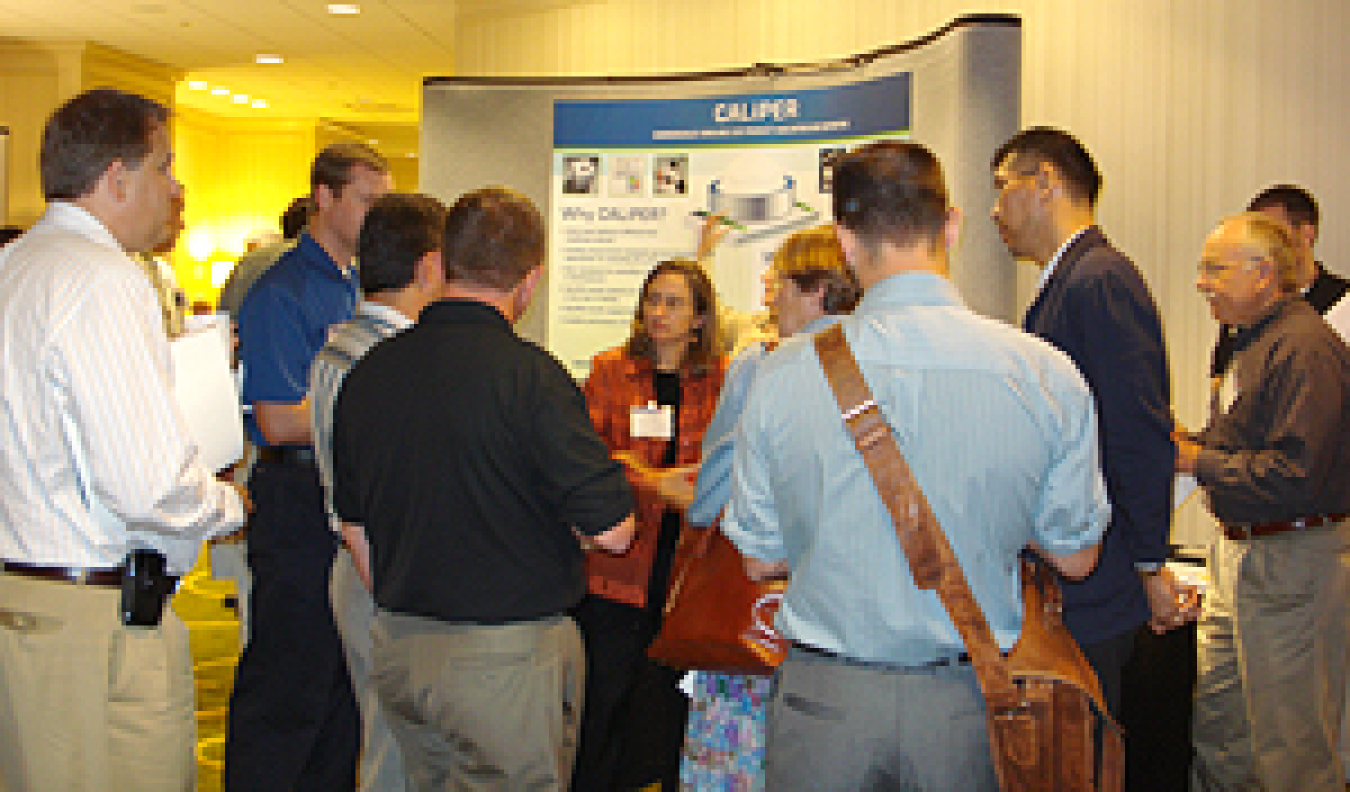 Photo of a group of people standing in an exhibit hall in front of a display marked CALiPER.