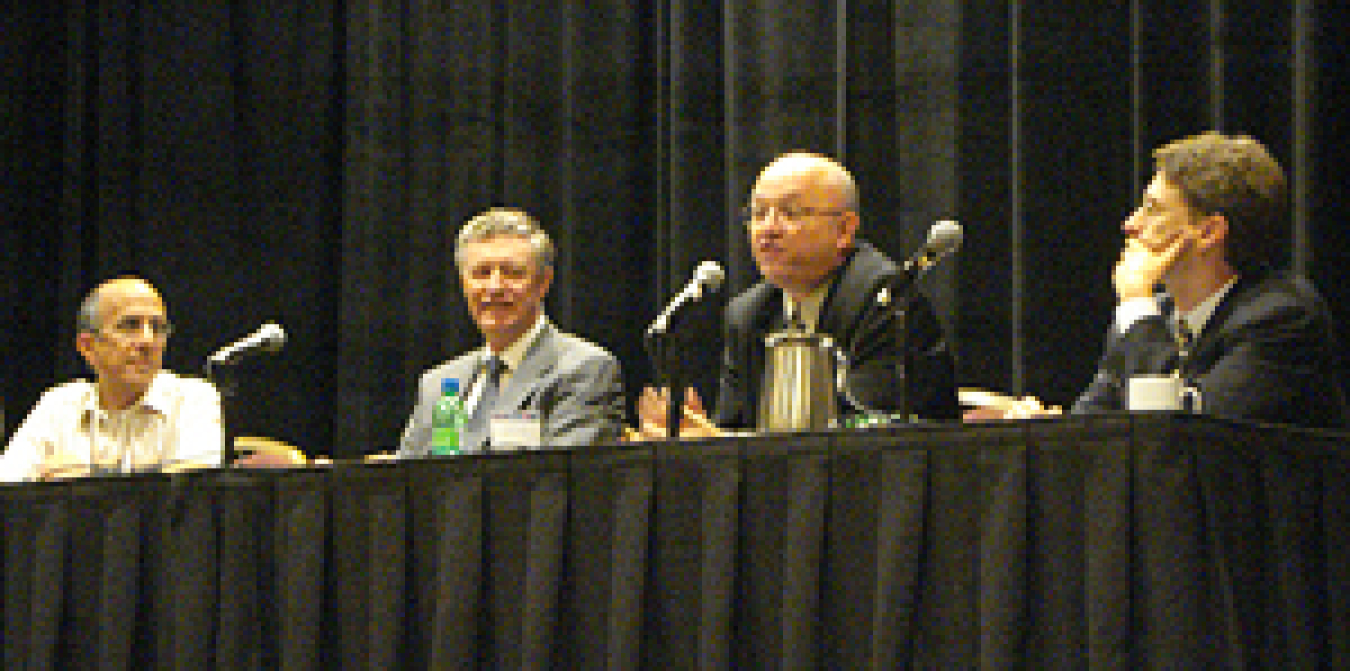 Photo of four men seated behind a long table with microphones on it, with one of the men speaking into the microphone in front of him.