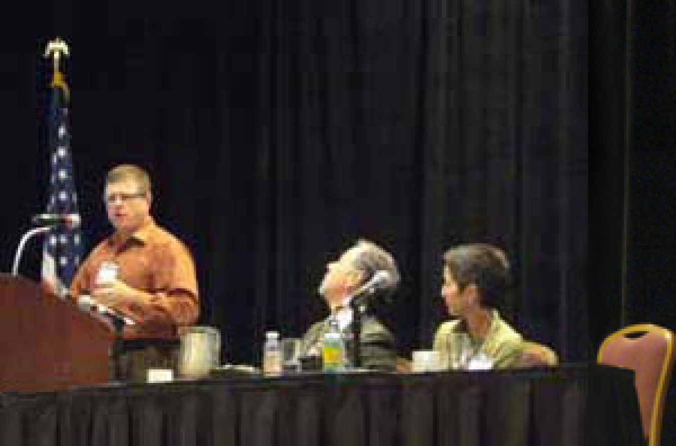 Photo of a man and a woman seated behind a long table. To their left are an American flag and a man at a podium speaking into a microphone.