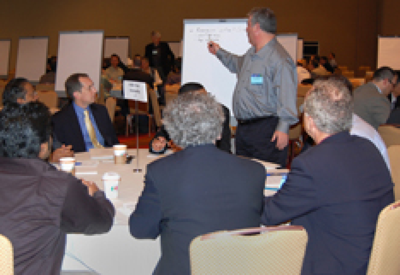 Photo of a group of men sitting at a table, looking toward a man who stands in front of an easel, pointing at a piece of paper.