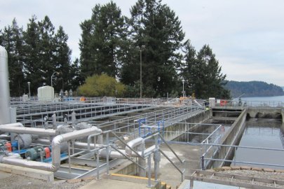 Image shows the tanks, pipes, and water holding areas of a wastewater treatment plan. Trees, shrubs, and a body of water are in the background.