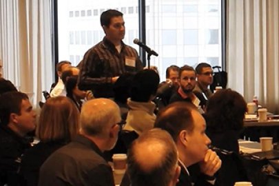 photo of a man standing at a microphone with a seated crowd of people around him.