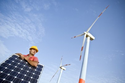A male construction worker stands by a solar panel with a wind turbine in the background. 