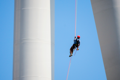 Worker hanging from cable between wind turbine rotor and tower with blue sky behind him.