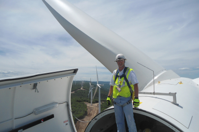 Man in a harness standing on top of an open wind turbine with other wind turbines in the distance.