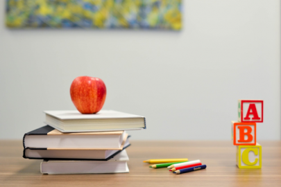 Photo of stacks of books on a table, with an apple on top, next to a few assorted-color pencils, and three building blocks with "A," "B" and "C" on them.