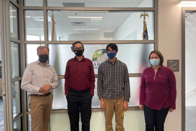 AEO Director George Roe stands to the left of three interns in front of the office wall. All three are wearing masks. 