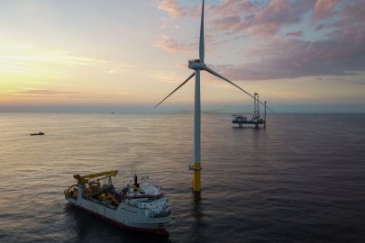 An offshore wind turbine in a calm ocean with a service vessel nearby; a platform is in the distance, erecting another wind turbine. The sun is rising (or setting) in the distance behind light clouds.