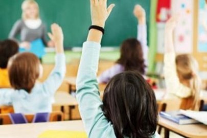 A school classroom, with backs of child students shown with raised hands and a teacher facing them blurry in the background.