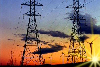Large electrical grid towers stand alongside wind turbines, with the sun setting behind them.
