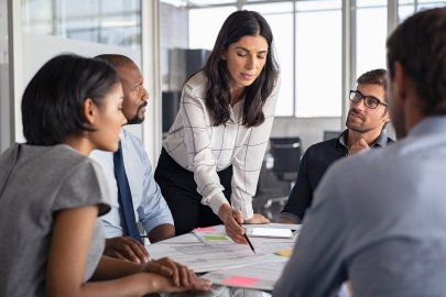 Group of people sitting at a table in a meeting, with a woman standing and pointing to a document on the table.
