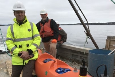 Two men in hard hats and stand next to a kayak on the deck of a boat on the water. One man is wearing a life jacket, and one is wearing a neon yellow jacket.