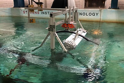 Two metal devices face each other from opposite ends of a wave tank that looks like an indoor swimming pool. Stevens Institute of Technology is written on the inside of the tank wall just above the water surface. Hanging over the water, a network of blue metal beams support a pair of electronic boxes on a track.