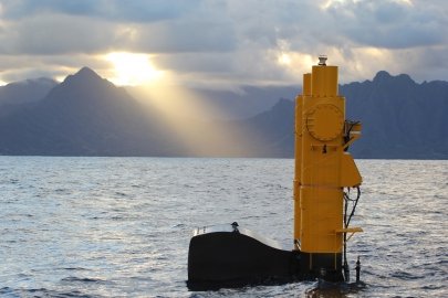A marine energy device floats on the surface of the ocean. Mountains and cloudy skies are visible in the background. The sun shines through a break in the clouds.