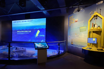 A museum exhibit with models of marine energy devices. A large screen at the center of the exhibit displays animations of different marine energy technologies.