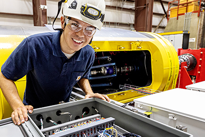 : A researcher in a hard hat leans over a hatch containing electronics and wires.