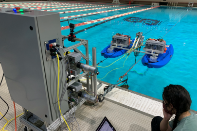 A photo of a researcher sitting next to a pool with a computer while two blue floating structures holding electronics pull the kite through the water.