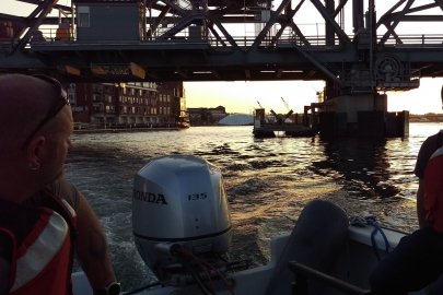 A person driving a motorboat in a narrow channel near a metal bridge at dusk.
