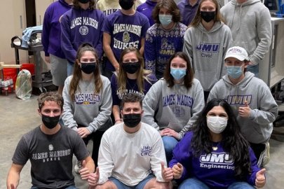 Sixteen students wearing medical masks in a research laboratory. Some are seated; others stand behind them. Most of the people are wearing shirts with the James Madison University logo.