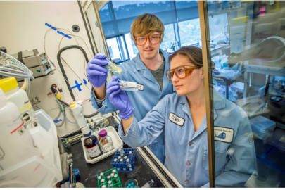 Two people in blue labcoats and safety goggles.