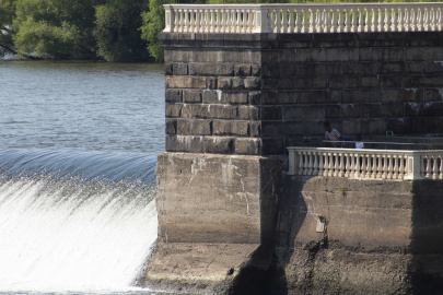A stone wall with a waterway behind it and a small waterfall as the water passes from the waterway past the stone structure 